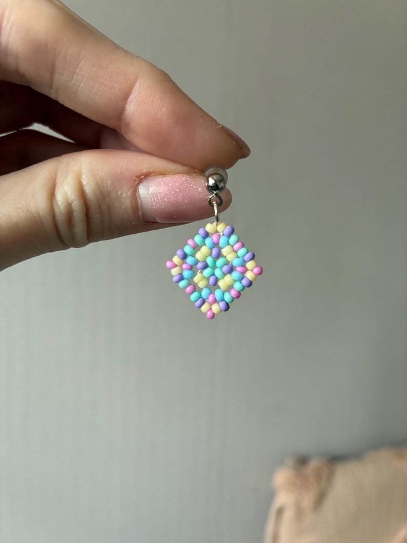 May include: A single beaded earring with a silver post. The earring features a diamond-shaped design made of small, colorful beads in pastel shades of pink, yellow, blue, and purple. The earring is held by a hand.