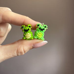 Puede incluir: Dos figuras de ranas de cuentas en tonos verdes, con ojos negros y mejillas rosas. Las ranas son peque&ntilde;as, con una textura detallada de cuentas. Una rana es de color verde claro y la otra de color verde oscuro. Las ranas se sostienen entre los dedos.