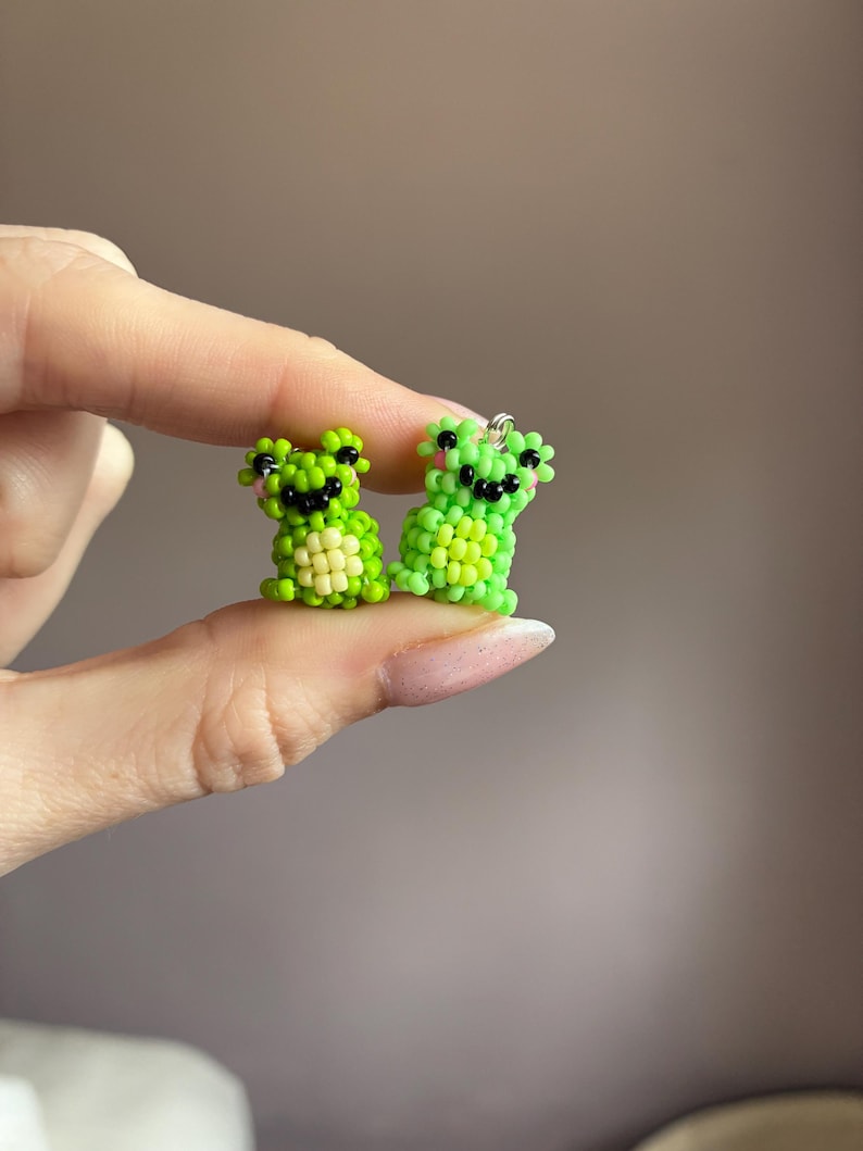 Puede incluir: Dos figuras de ranas de cuentas en tonos verdes, con ojos negros y mejillas rosas. Las ranas son peque&ntilde;as, con una textura detallada de cuentas. Una rana es de color verde claro y la otra de color verde oscuro. Las ranas se sostienen entre los dedos.