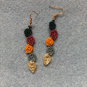 Pu&ograve; includere: Un paio di orecchini fatti a mano con perline e ganci dorati. Gli orecchini presentano perline a forma di foglia in vari colori, tra cui verde scuro, bordeaux, arancione, argento e oro. Le perline sono disposte in una linea verticale.