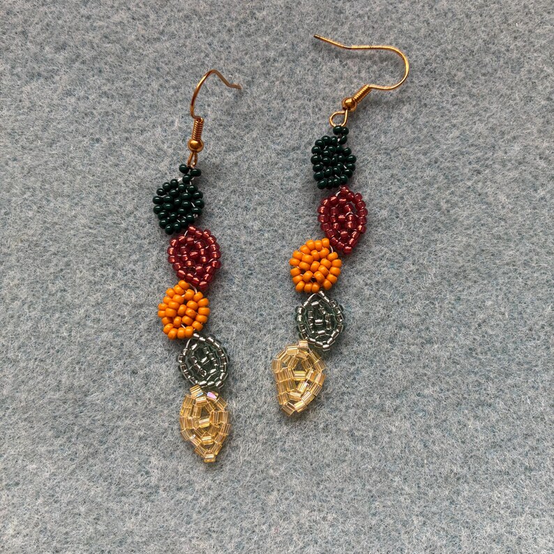 Pu&ograve; includere: Un paio di orecchini fatti a mano con perline e ganci dorati. Gli orecchini presentano perline a forma di foglia in vari colori, tra cui verde scuro, bordeaux, arancione, argento e oro. Le perline sono disposte in una linea verticale.