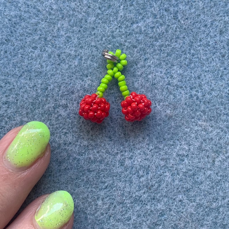 May include: A beaded cherry charm with bright red cherries and a green stem. The charm is made of small beads and has a silver-colored ring at the top for attaching to a keychain or other item. The background is a textured blue surface.