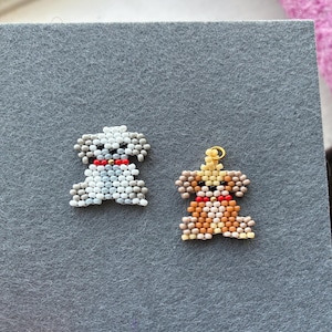 May include: Two beaded dog-shaped charms. One is white and light blue, the other is brown, tan, and yellow. Both have red collars. The charms are on a gray felt surface.