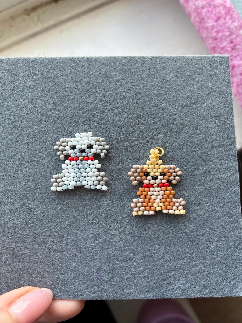 May include: Two beaded dog-shaped charms. One is white and light blue, the other is brown, tan, and yellow. Both have red collars. The charms are on a gray felt surface.