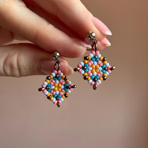 May include: A pair of colorful beaded earrings. Each earring features a square design with a mix of pink, blue, orange, and black beads. The earrings have a silver-colored post and a small gold-colored accent.