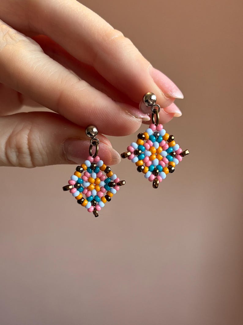 May include: A pair of colorful beaded earrings. Each earring features a square design with a mix of pink, blue, orange, and black beads. The earrings have a silver-colored post and a small gold-colored accent.