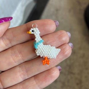 Puede incluir: Un charm de ganso hecho a mano con cuentas blancas, con una bufanda azul, patas naranjas y un pico amarillo y negro. El charm tiene un lazo plateado en la parte superior para colgar. Un accesorio caprichoso.