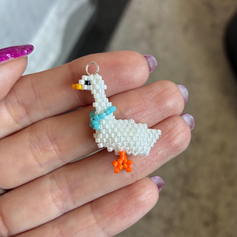 Puede incluir: Un charm de ganso hecho a mano con cuentas blancas, con una bufanda azul, patas naranjas y un pico amarillo y negro. El charm tiene un lazo plateado en la parte superior para colgar. Un accesorio caprichoso.