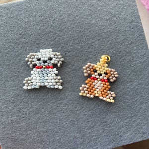 May include: Two beaded dog charms. One is white and gray, the other is brown and tan. Both have red collars. The charms are on a gray felt surface. The charms are small and detailed.