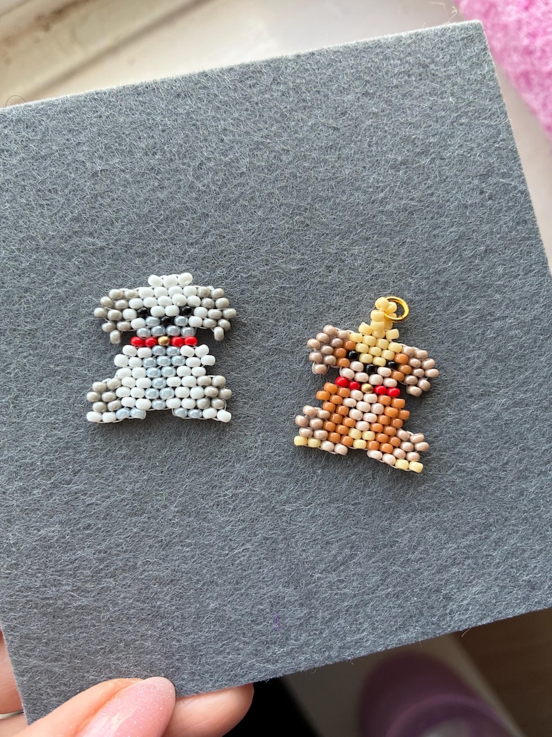 May include: Two beaded dog charms. One is white and gray, the other is brown and tan. Both have red collars. The charms are on a gray felt surface. The charms are small and detailed.
