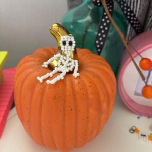 May include: An orange pumpkin with a gold stem, topped with a beaded white skeleton. The pumpkin is a decorative item, likely for Halloween or fall. Other decorative items are in the background.