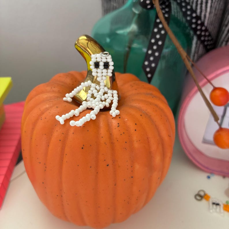 May include: An orange pumpkin with a gold stem, topped with a beaded white skeleton. The pumpkin is a decorative item, likely for Halloween or fall. Other decorative items are in the background.