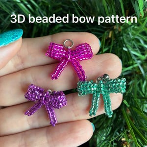 Könnte beinhalten: Drei Perlen-Schmuckanhänger in Form von Schleifen in Lila, Rosa und Grün. Die Schleifen sind aus kleinen Perlen gefertigt und haben ein 3D-Design. Der Text "3D beaded bow pattern" ist oben im Bild zu sehen.