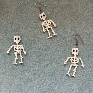 May include: Three white beaded skeleton earrings with black bead eyes and silver-colored ear wires. The earrings are on a light blue background. The skeleton earrings are a Halloween or gothic theme.
