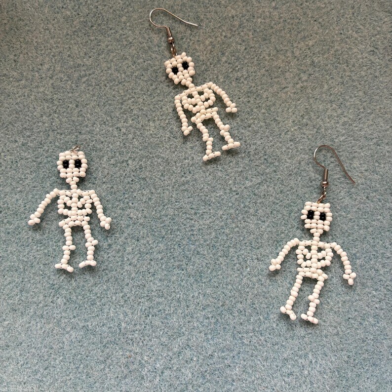 May include: Three white beaded skeleton earrings with black bead eyes and silver-colored ear wires. The earrings are on a light blue background. The skeleton earrings are a Halloween or gothic theme.