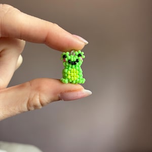 Puede incluir: Un peque&ntilde;o amuleto de rana con cuentas en tonos verdes y amarillos. La rana tiene ojos negros y una boca sonriente, con detalles en rosa. El amuleto est&aacute; sujeto por un peque&ntilde;o lazo de metal.