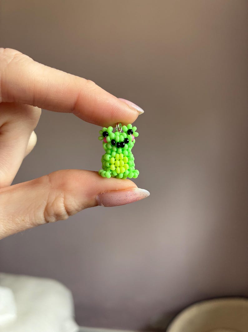 Puede incluir: Un peque&ntilde;o amuleto de rana con cuentas en tonos verdes y amarillos. La rana tiene ojos negros y una boca sonriente, con detalles en rosa. El amuleto est&aacute; sujeto por un peque&ntilde;o lazo de metal.