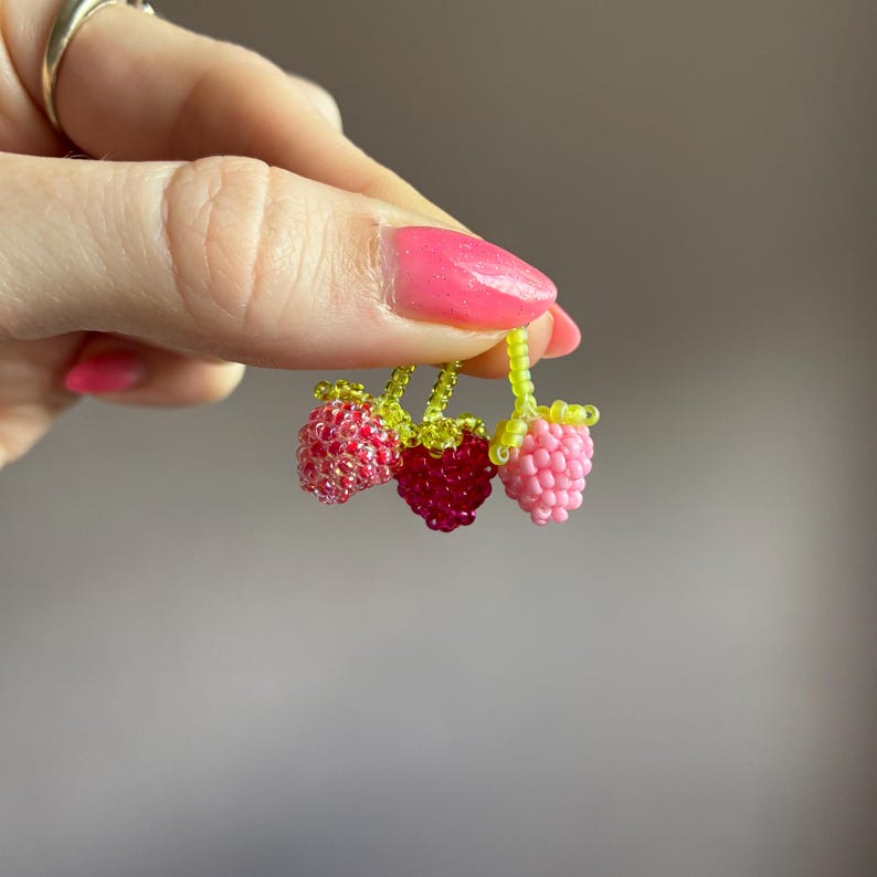 May include: A cluster of three small berry-shaped charms crafted from tiny glass beads. The charms feature a light pink berry, a deep magenta berry, and a variegated red and pink berry, each topped with a bright lime green beaded stem.