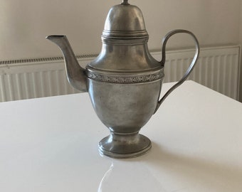 Op de afbeelding: Een zilveren theepot met een decoratief ontwerp rond het midden. De theepot heeft een deksel met een knop bovenop.