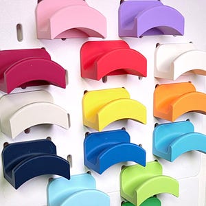 May include: A collection of colorful, curved, plastic hat holders in various shades, including pink, purple, red, white, yellow, orange, blue, green, and brown. The holders are mounted on white pegboards, offering a vibrant display.