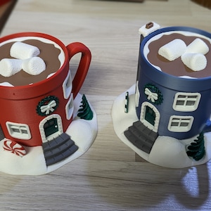 Puede incluir: Dos tazas decorativas con forma de casas, una roja y otra azul, cada una llena de chocolate caliente y malvaviscos. Las tazas tienen ventanas, puertas y chimeneas, y se asientan sobre una base nevada con un árbol de Navidad y un bastón de caramelo.