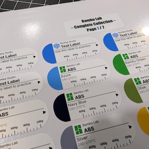 Puede incluir: Una hoja de etiquetas adhesivas blancas para filamento de impresora 3D, con varios colores y texto que incluye Bambu Lab, ABS y marcas de peso de 200g a 800g. Las etiquetas est&aacute;n organizadas en filas con pesta&ntilde;as de colores y logotipos de marca.