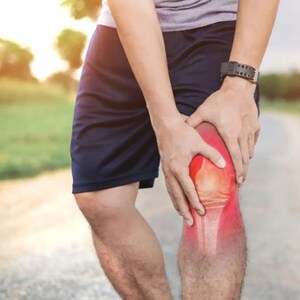 Könnte beinhalten: Eine Person hält ihr Knie, mit einem rot hervorgehobenen Bereich, der Schmerzen anzeigt. Die Person trägt ein graues Hemd, schwarze Shorts und eine schwarze Uhr. Der Hintergrund zeigt eine verschwommene Umgebung im Freien.