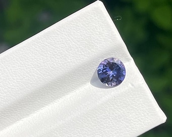May include: A round, faceted gemstone displaying a gradient of violet and blue hues. The stone is set into a white, textured display card with a visible crease, highlighting the reflective surfaces and internal light play of the circular gem.