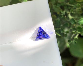 May include: A loose, triangular-cut gemstone with a vibrant blue and violet color palette. The stone features a faceted surface that reflects light, creating internal patterns of deep blue and purple hues set against a white display surface.