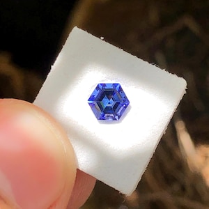 Könnte beinhalten: Ein facettierter, sechseckiger Edelstein in tiefblau. Der Stein wird auf einem weißen Quadrat gehalten, was seinen komplizierten Schliff und seine Brillanz hervorhebt. Die Facetten des Steins reflektieren das Licht.