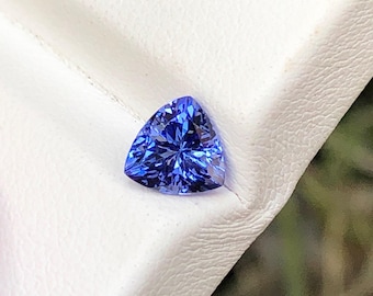 May include: A loose, trillion-cut gemstone with a vibrant, deep blue color and a faceted surface that reflects light. The stone has a triangular shape with sharp, pointed corners and is displayed against a white, textured surface.