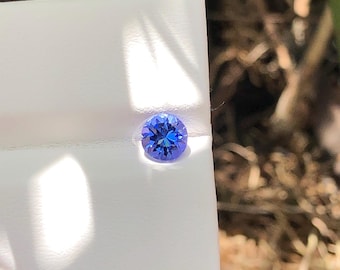 May include: A round, faceted gemstone with a vibrant blue and violet hue. The stone is set into a white display card, showcasing its brilliant cut and reflective surface. The gemstone displays a clear, transparent quality with deep color saturation.