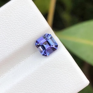 Puede incluir: Una gema tallada cuadrada, de color azul-violeta intenso. La gema está colocada sobre una superficie blanca texturizada, con un fondo verde borroso. Las facetas de la gema reflejan la luz. Ideal para joyería.