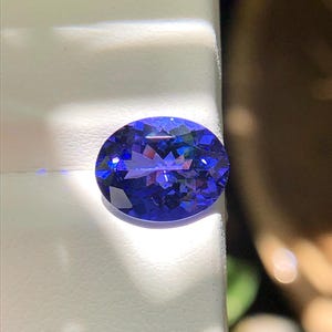 Può includere: Una gemma sfaccettata di forma ovale con una tonalità blu intenso. La gemma è posta su uno sfondo bianco, con la luce che si riflette sulle sue numerose sfaccettature. La gemma sembra essere una tanzanite.