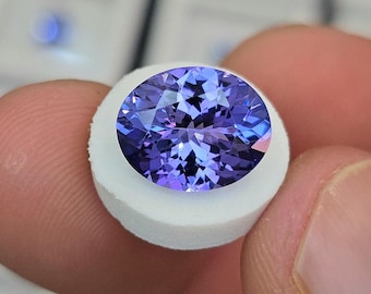Puede incluir: Una gema ovalada facetada de color azul-violeta intenso, colocada sobre una base blanca. La estructura interna de la gema refleja la luz, creando un efecto brillante. El fondo está desenfocado, resaltando la gema.
