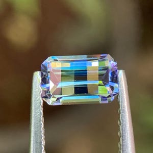 Puede incluir: Una gema de corte rectangular sujeta con pinzas plateadas. La gema muestra un espectro de colores, incluyendo azules, verdes y amarillos, con un efecto iridiscente brillante. Las facetas de la piedra reflejan la luz.
