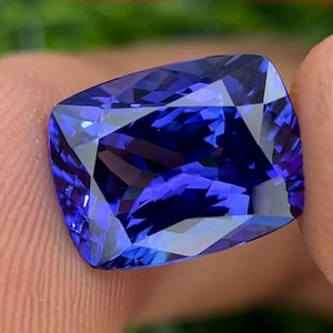 May include: A cushion-cut, faceted gemstone with a deep blue hue. The gem's facets reflect light, creating a brilliant display of color and sparkle. The stone is held between fingers, showcasing its size and shape.
