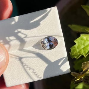 May include: An oval-cut gemstone with a pale, translucent appearance and subtle light-reflecting facets. The stone is set against a white, textured square display card, catching natural light that creates distinct shadows across the surface.