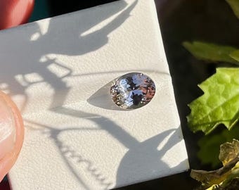 Puede incluir: Una piedra preciosa de corte ovalado con una apariencia pálida y translúcida y facetas sutiles que reflejan la luz. La piedra está colocada sobre una tarjeta de presentación cuadrada de color blanco y texturizada, captando la luz natural.