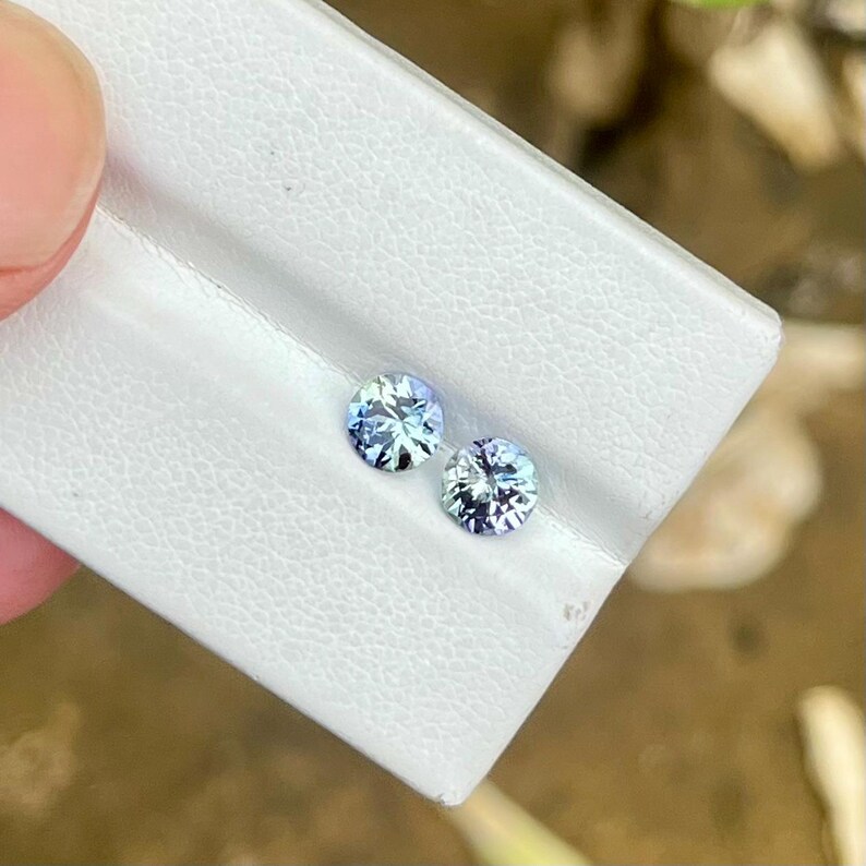 May include: Two round, faceted gemstones with a light blue and purple color. The gems are set on a white surface and appear to be held by a person's fingers. The gems are likely for jewelry making.