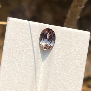 May include: An oval-cut gemstone with a light brown and champagne hue, featuring a faceted surface that reflects light. The stone is set against a white textured display card, showcasing its translucent quality and polished finish.