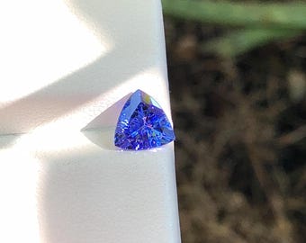 May include: A loose, trillion-cut gemstone with a vibrant blue and violet hue. The stone features a faceted surface that reflects light, creating internal sparkle and depth. It is positioned on a white display card with a smooth, matte texture.