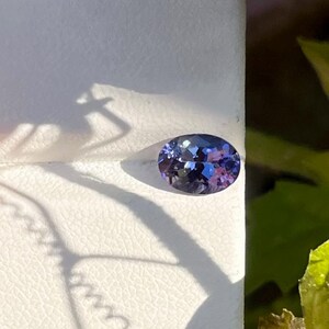 May include: An oval-cut gemstone displaying a vibrant mix of deep violet, blue, and purple hues. The stone features a faceted surface that reflects light, creating internal sparkles and color shifts across its polished, translucent, and rounded shape.