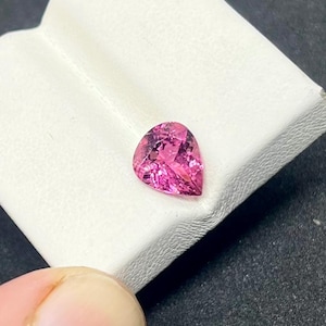 Puede incluir: Una gema rosa facetada en forma de pera. La gema es de un tono rosa vibrante, con múltiples facetas que reflejan la luz. La gema está sobre una superficie blanca.