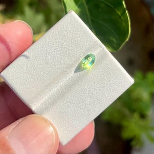 Pode incluir: Uma pequena pedra preciosa de corte oval com um tom vibrante de verde-lima e amarelo, apresentando uma superfície facetada que reflete a luz. A pedra está montada num cartão de exposição quadrado branco com uma textura granulada.