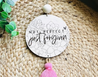 May include: White round wooden sign with a black outline of succulents and the text "Not perfect just forgiven" with a pink heart. The sign is hanging from a white bead and has a pink tassel.