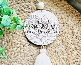 May include: A wooden round sign with a white background and black text that reads "created for a purpose" with a heart symbol. The sign is decorated with a black and white line drawing of succulents. The sign is hanging from a white cord with a white wooden bead. The sign is hanging from a brown woven surface. The sign is attached to a three-tiered tassel with white, beige, and brown colors.