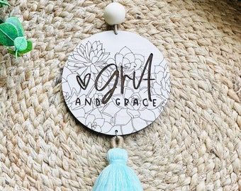 May include: A round wooden ornament with the words "GNA AND GRACE" and a heart symbol, surrounded by a floral design. It has a light blue tassel and a wooden bead for hanging. The background is a woven, natural fiber.