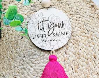 May include: A round wooden sign with a floral design and the text "Let your light shine Matthew 5:16". The sign is attached to a pink tassel.