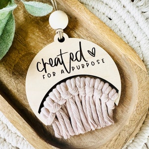 May include: A wooden half-moon shaped ornament with the text "created for a purpose" and a small heart. The ornament has a pink macrame tassel hanging from it.